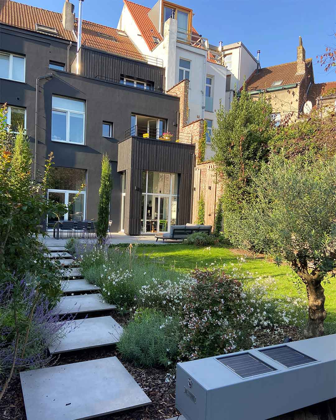 Jardin moderne avec allée en dalles de béton, massifs fleuris et terrasse ensoleillée au pied d’une maison rénovée.